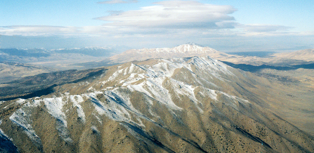 [The airplane wing was edited out of this image. It contains brown mountains with many small sections with white covering(snow). This image is much closer to the tops of the mountains than the prior image. The ground is brown on the mountains and on all sides of them.]